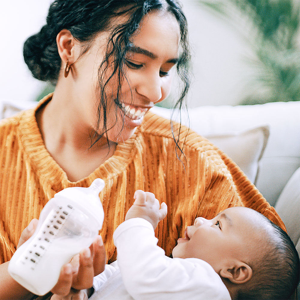 A mother sits on the couch, gently bottle-feeding her newborn with infant formula.