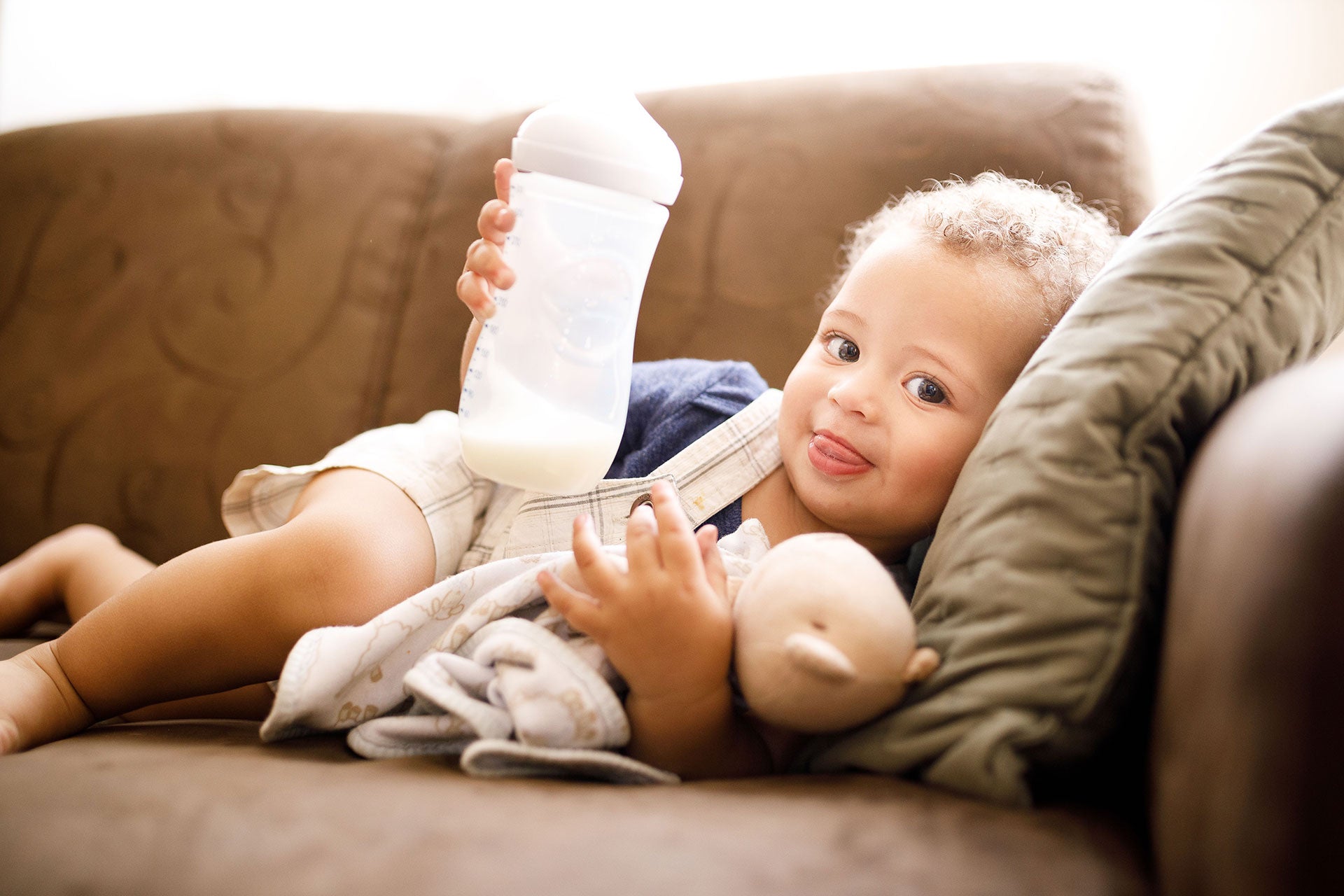 A baby sticks his tongue out as he lies on the couch holding a stuffed teddy in one hand and a bottle of formula in the other.