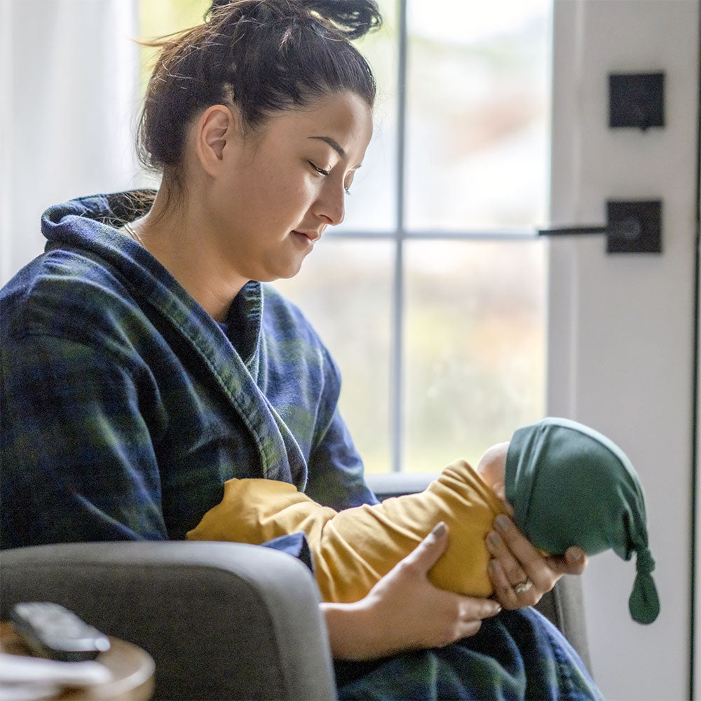 A mom in a housecoat looks down affectionately as she holds a swaddled baby.