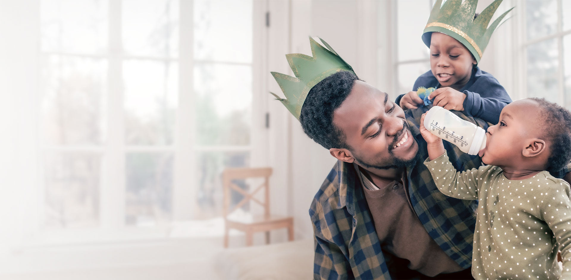 Un papa jouant joyeusement sur le plancher avec ses deux tout-petits, souriant tandis que l'un est dans son dos et que l'autre boit à la préparation.