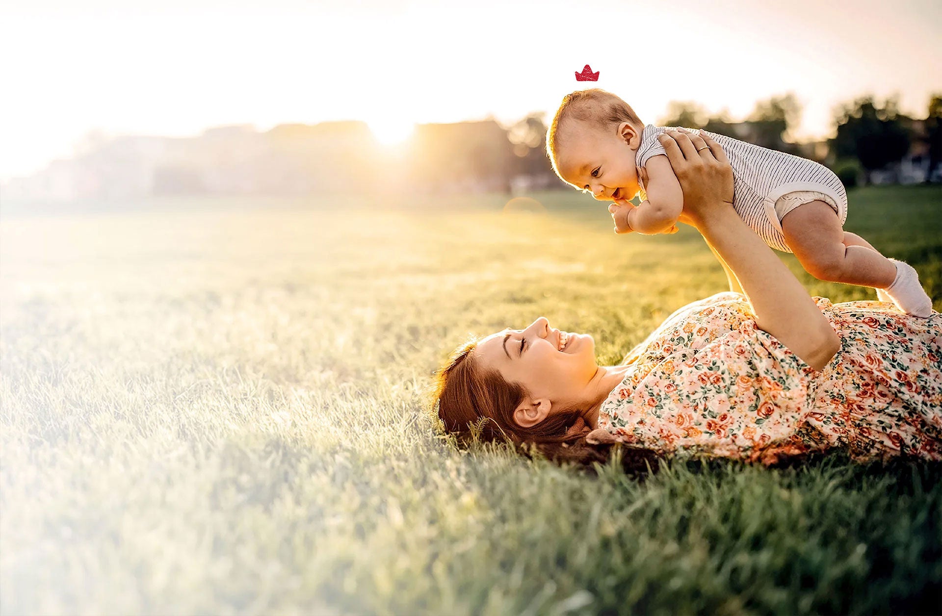 A mom lies on her back in the grass, holding a baby wearing a Niuriss crown to represent our formulas.