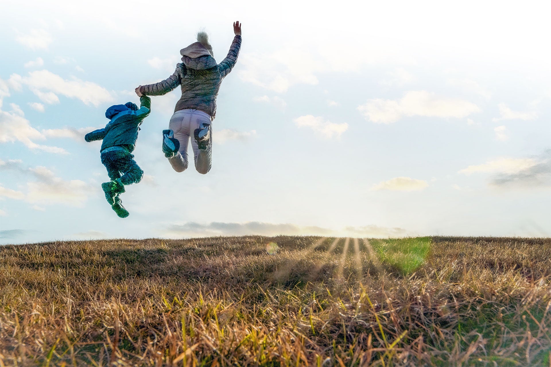 A parent and toddler hold hands on an outdoor walk, jumping excitedly while facing away from the camera.