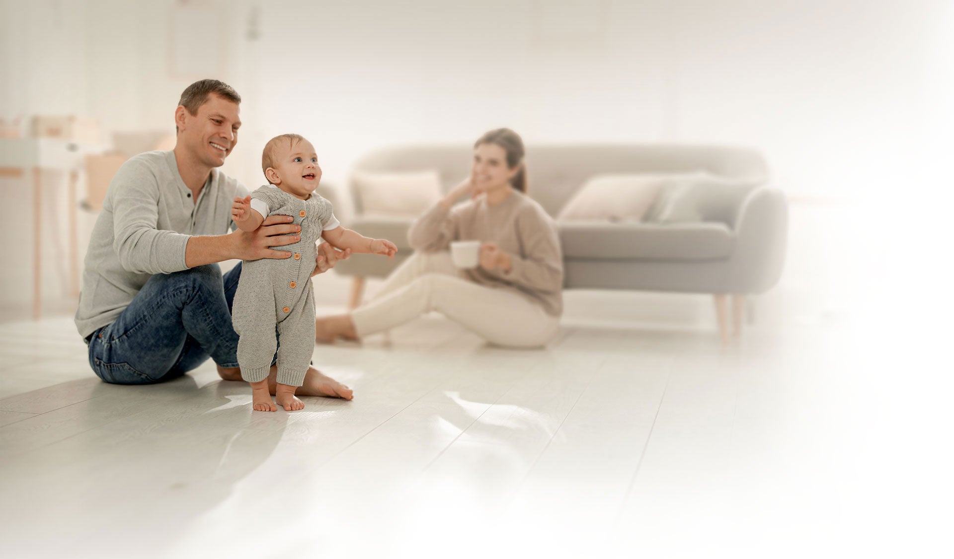 A baby smiles as he stands, supported by his father who sits on the floor. His mother smiles in the background.