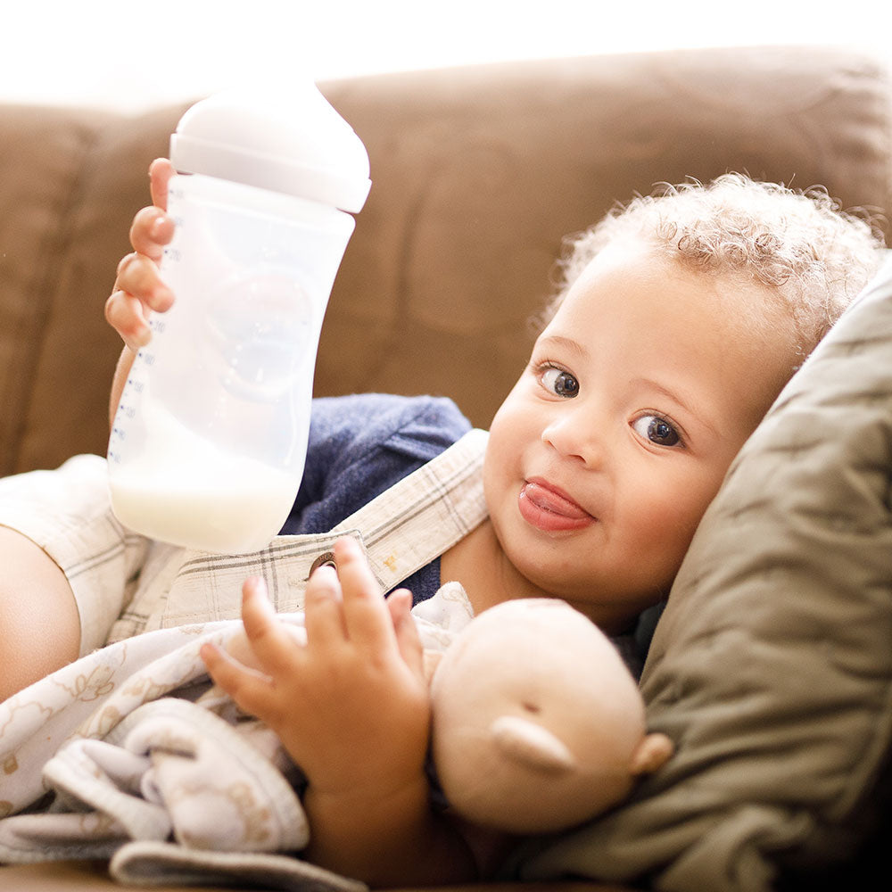 A smiling toddler on the couch, holding a stuffed bear in one hand and toddler formula in the other.